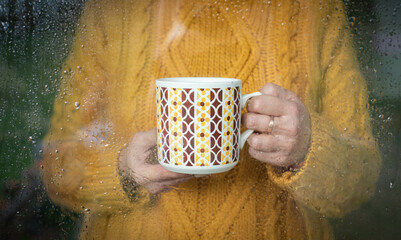Woman in mustard yellow sweater holding a retro porcelain mug with colorful pattern, behind the raindrops covered window