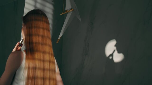 Rear view: a girl with long red hair stands and paints her lips with lipstick, the profile of a beautiful face is reflected on the wall. The play of light and shadow, the concept of beauty. Slow mo,4K