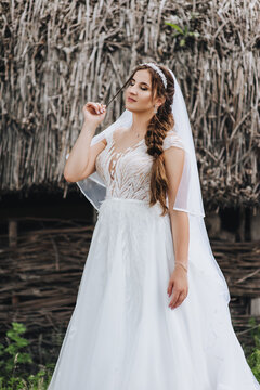 A beautiful, sweet, tender bride in a white dress with a veil and a diadem on her head, standing in the village against the background of a hut with a straw. Wedding photography close-up, portrait.