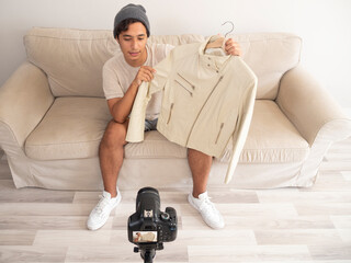 Hispanic young man creating clothing content for social networks with a camera on a tripod, sitting on a piece of furniture
