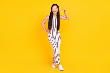Full length body size photo girl smiling in striped overall showing v-sign gesture isolated vibrant yellow color background