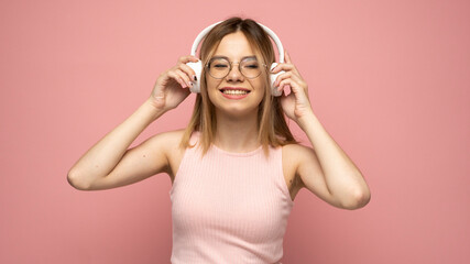 Obraz na płótnie Canvas Beautiful attractive young blond woman wearing pink t-shirt and glasses in white headphones listening music and smiling on pink background in studio. Relaxing and enjoying. Lifestyle.