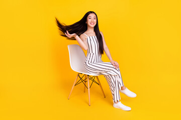 Full length body size photo young girl sitting on chair wearing casual overall smiling throwing hair isolated vivid yellow color background