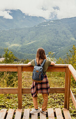 Fototapeta na wymiar Rear view of young blonde woman with backpack and plaid shirt looking at view of Caucasian mountains and cloudy sky standing on wooden viewpoint, hiking active healthy lifestyle nature
