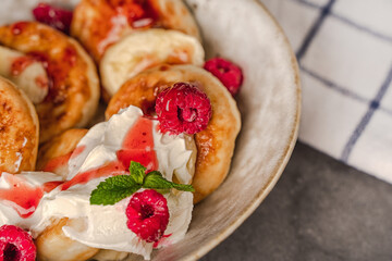 Delicious cottage cheese pancakes in a ceramic plate with yogurt and raspberries on a dark background with a towel.