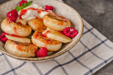 Obraz na płótnie Canvas Delicious cottage cheese pancakes in a ceramic plate with yogurt and raspberries on a dark background with a towel.