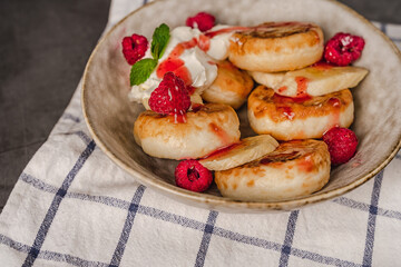 Delicious cottage cheese pancakes in a ceramic plate with yogurt and raspberries on a dark background with a towel.