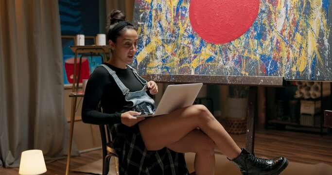 A tired artist, after finishing new painting, sits at canvas with a laptop on lap, creating sale auctions, taking a video call through her webcam, waving to a friend, talking, gossiping, laughing