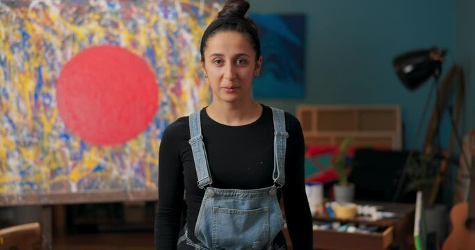 Tired artist dressed in dungarees stands in front of abstract painting with big red dot, masterpiece on display, painting studio, girl rubs forehead holding brush smiles at camera
