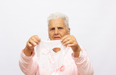 Portrait of senior woman in bath towel taking off rejuvenating cotton face mask on light studio background. Lovely mature lady using anti-aging cosmetic product how to use cosmetic mask