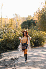 Portrait of a happy cheerful young woman in fashionable casual clothes and a stylish hat walking drinking coffee enjoying solitude in a fall park in nature by the water in autumn, selective focus