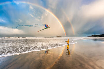 Girl in yellow coat flying kite on beach with double rainbow