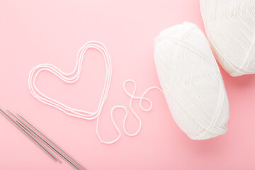Heart shape created from white wool, metal needles and clews on light pink table background. Pastel...