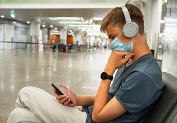 Serious young man teenager 15-18 years old typing a message on the phone while sitting in the airport lounge. Young student in casual clothes and a protective face mask using a smartphone.