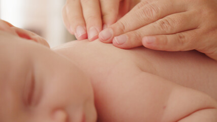 Mother doing massage on her healthy sleeping infant baby. Small caucasian newborn resting and laying on his belly while his mother is performing a massage for his small back and developing muscles. .