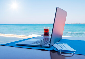 Mobile office on the beach with turquoise sea open laptop, blue sky, Turkish tea and protective face mask.