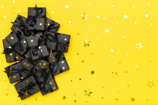 Black gift boxes with black ribbons and golden glitter stars confetti sparkles on bright rich yellow background. Black Friday sale flat lay, top view, copy space. Holiday, festive, party backdrop.
