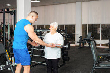 pensioner, an elderly woman performs an exercise with a barbell in gym. coach helps and shows. Healthy sports lifestyle, senior concept. woman with gray hair is recovering from injury.