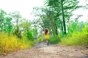 Happy little girl with long hair in a yellow dress  running  along path on meadow with sunset