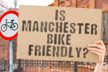 The question " Is Manchester bike friendly? " on a banner in men's hand with blurred background. Transportation. Zero waste. Bicycle lane. Streets. City. Safety. Insecure. Road signs. Dangerous