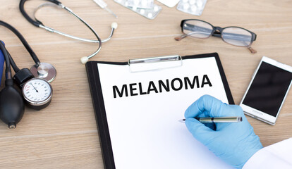 text MELANOMA written on paper by the hand of a doctor, on the table next to stethoscope, glasses, pills, medical concept.