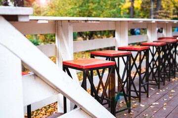Empty street cafe with a row of red bar stools in the park against a background of golden autumn foliage and a white fence.