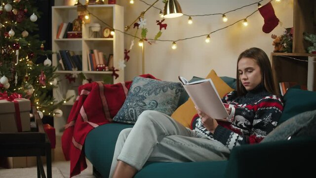 Woman reading book and drinking tea close-up. Young student sitting on sofa in decorated living room. Christmas holidays concept. Female person resting at home, leisure, new year time. 