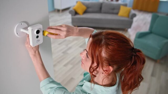 mounting wireless cam on screws for home security system, young woman installing surveillance camera on wall and then picks up a tablet