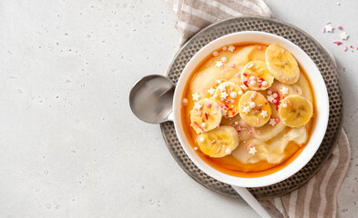 Semolina porridge with syrup and banana. Sweet breakfast, baby food. Semolina porridge in a white bowl on a gray concrete background top view. Copy space for text