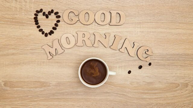 Coffee. A cup of black coffee is moving on a wooden table. Later, the inscription Good morning and a heart of coffee beans appears. Background, flat lay, top view. Stop motion.