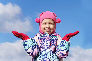 Cute little girl on a background of blue sky with raised hands. Pink clothes. Child outside in winter in cold weather