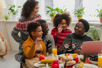 cheerful african american girl waving hand during video call near family