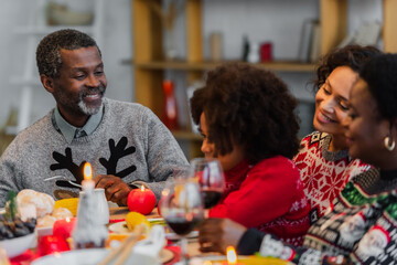 happy african american man looking at granddaughter near blurred family on christmas dinner