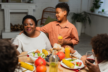 happy african american boy with orange juice hugging grandmother during thanksgiving dinner with blurred family