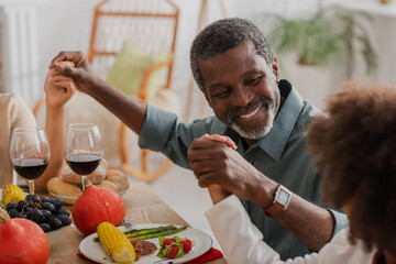 cheerful african american man holding hands with family while praying on thanksgiving dinner