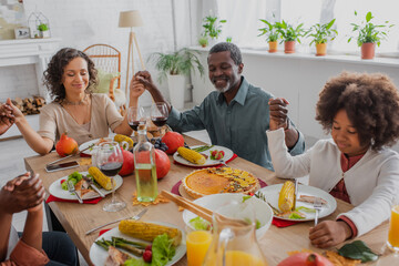 happy african american family holding hands while praying before thanksgiving dinner