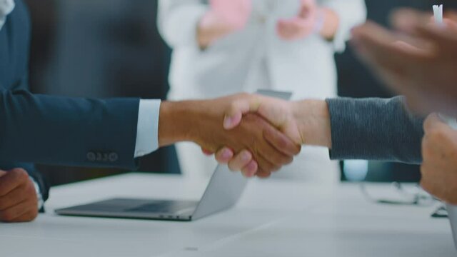 Cropped view of handshake above desk. Colleagues around clapping hands. Selective focus. Modern open space office interior.