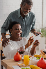 African american woman using smartphone near husband with wine and thanksgiving dinner