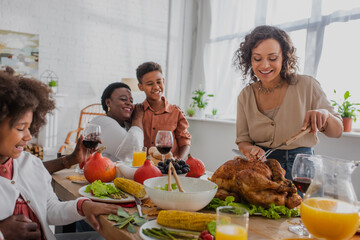 Smiling african american woman cutting turkey near family and thanksgiving dinner