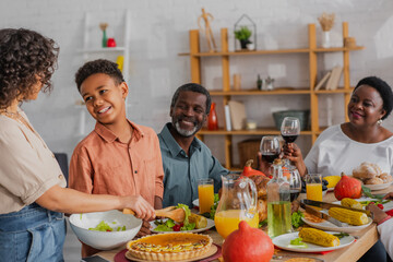 African american boy looking at mother pouring salad during thanksgiving dinner with grandparents