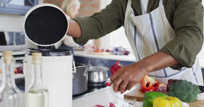 African american senior man wearing apron putting vegetable peelings in a compost bin in the kitchen