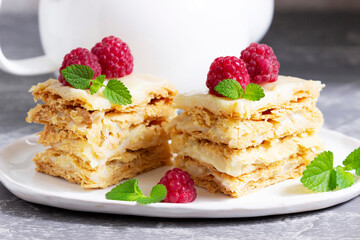 Pieces of Napoleon cake decorated with raspberries and lemon balm leaves on a light background.