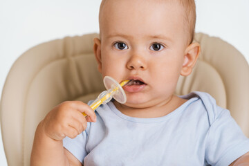 A one-year-old child is sitting on a chair with a toothbrush in his hands. Brushing teeth by a...