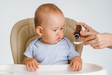 A one-year-old child sitting on a chair watches as his mother dials the dosage of medicine into a...