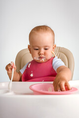 A baby 12-15 months old sits on a feeding chair and reaches into a plate of food with his hand,...