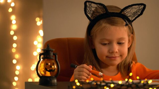Little girl sits in dark at table with halloween pumpkin lamp and paints orange marker on background of shining golden blurred bokeh.