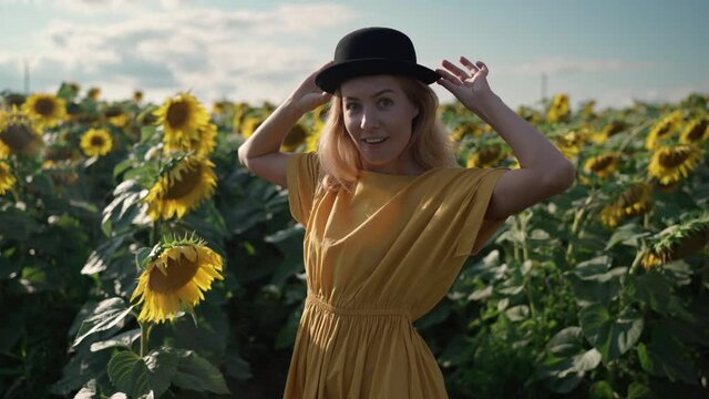 beautiful redhead caucasian woman in a long yellow dress and black hat on a sunflower field on a sunny day at a photo shoot taking pictures by phone, posing, smiling, indulging