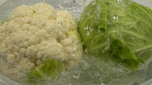 Running water pours onto the white cabbage and the cauliflower head lying in a container with water. The washing vegetables  before cooking