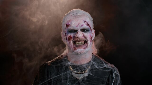 Zombie man with makeup with fake wounds scars and white contact lenses looking at camera and clicks his teeth, trying to scare in dark room. Sinister dead guy. Halloween, filming, staging concept