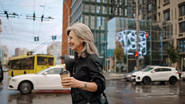 Adult gray-haired woman in glasses walks along a city street with a folder and coffee in her hands. Business lady walk. Slow motion